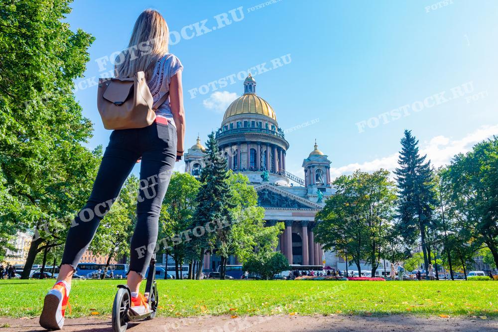 На фотографии запечатлен момент, когда женщина на самокате любуется Исаакиевским собором в Санкт-Петербурге. Она стоит спиной к камере, и это создаёт ощущение её участия в городской жизни, её движения и динамики. Здание собора возвышается вдалеке, окружённое зелёными деревьями и синим небом. Фотография может быть использована в туристических брошюрах, статьях о жизни в городе или как изображение для вдохновения на активный отдых и исследования городских пространств.